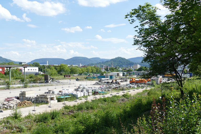 Das Bild zeigt den ein städtisches Baugebiet mit Baumaterialien und Baugeräten im Vordergrund, umgeben von grüner Vegetation, mit einem Stadtpanorama und Hügeln im Hintergrund.