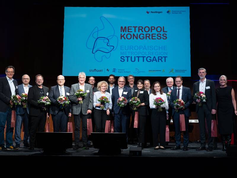 Eine Gruppe von Personen steht lächelnd auf einer Bühne und hält Blumensträuße, vor einem großen Bildschirm mit der Aufschrift "Metropol Kongress Europäische Metropolregion Stuttgart".