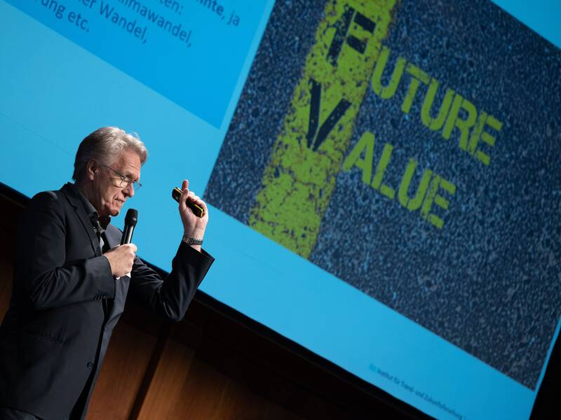 Eine Person hält ein Mikrofon und einen Präsentationszeiger in der Hand, während sie vor einem Bildschirm mit der Aufschrift "FUTURE VALUE" spricht.